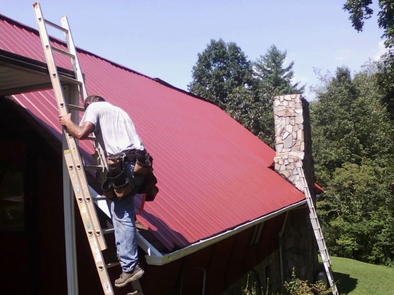 Roofer on a ladder beside a red metal roof and stone chimney — Soffit & Fascia Repair by Roofing Brothers in Graytown